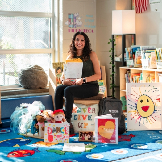 Sarah sits in her classroom holding a packet of letters and drawings from students and surrounded by puppets, books, pillows, and costumes that she uses in her class.